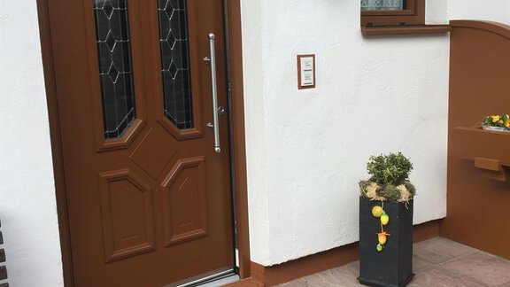 A brown entrance door with decorative windows and an exterior lamp. Next to it stands a flower pot with plants and a small decoration. - Appt. Schnöller - Sölden