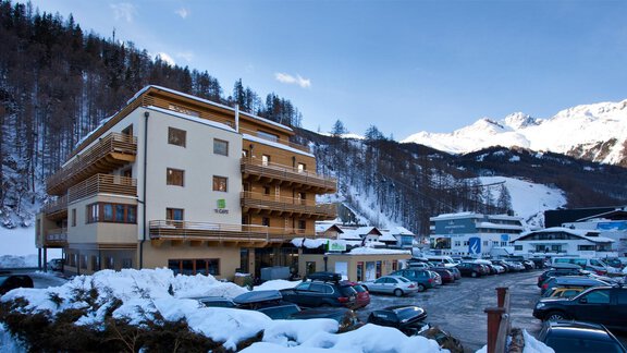 Ein modernes Hotel in einer schneebedeckten Berglandschaft. Im Vordergrund sind zahlreiche Autos auf einem Parkplatz zu sehen. - Bäckelar Wirt - Sölden