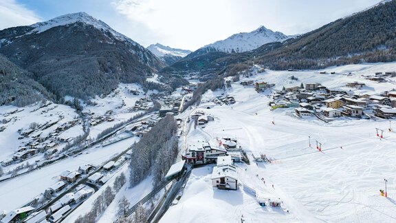 A winter mountain landscape with snow-covered hills and mountains. In the foreground, there is a ski resort with some buildings and slopes. - Belledonne Ski in & out Appartements - Sölden