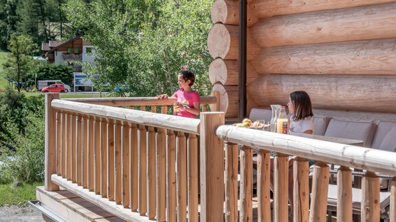 A wooden terrace with two people relaxing. In the background, there are trees and a rustic setting visible. - Chalet Resort Sölden - Sölden