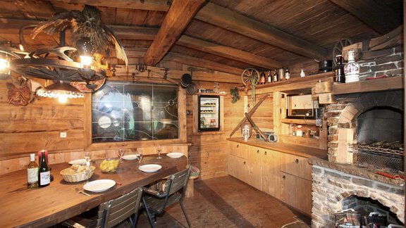 A cozy kitchen in rustic style with wooden walls and a large dining table. In the background, there is an open fireplace and a modern kitchenette. - Erbhof - Sölden