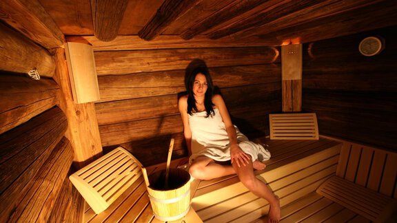 A woman is sitting relaxed in a wooden sauna. Next to her is a bucket of water. - Erbhof - Sölden