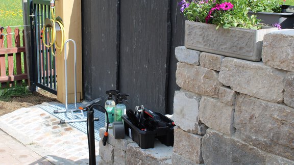 An area with a garden that features a stone facade and a water feature. In the foreground, there are cleaning tools and flowers in a flower box. - Ferienappartements Linserhof - Sölden