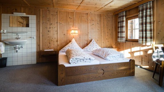 A cozy bedroom with a double bed and two large pillows. The walls are made of wood and there is a window with flowing curtains. - Ferienhaus Karlingerhof - Sölden