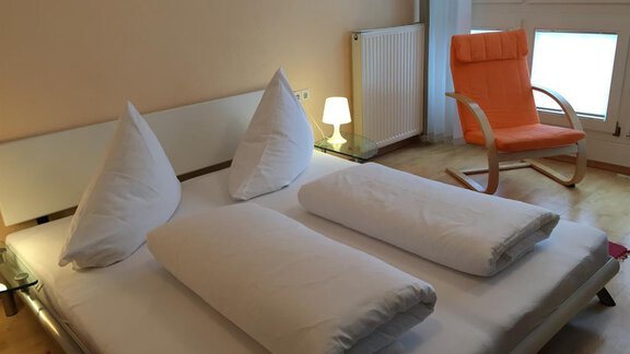 A simple bedroom with a double bed and two pillows. Next to the bed is a chair and a lamp in a bright room. - Ferienhaus Moidl - Sölden