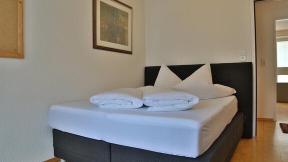 A simple bedroom with a double bed and fresh, white bedding. A picture hangs on the wall, and the door leads to another room. - Ferienhaus Moidl - Sölden