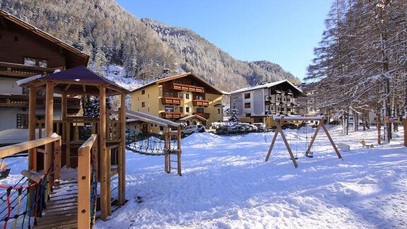 A playground in the snow with swings and play equipment. In the background, charming houses and snow-covered mountains can be seen. - Ferienhaus Waldele - Sölden