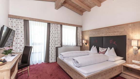 A cozy hotel room with wooden details and a large bed. The windows let in plenty of light and the carpet is red. - Garni Jaqueline - Sölden