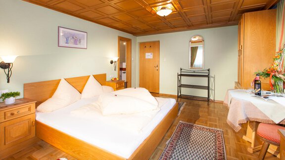 A cozy bedroom with a large double bed and a wooden shelf. The walls are painted in soft colors, and there is a table with a chair. - Gästehaus Kneisl - Sölden