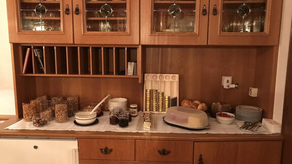 A wooden dresser with glasses and dishes. Various foods such as muesli, jam, and rolls are visible on the tabletop. - Gästehaus Martin - Sölden
