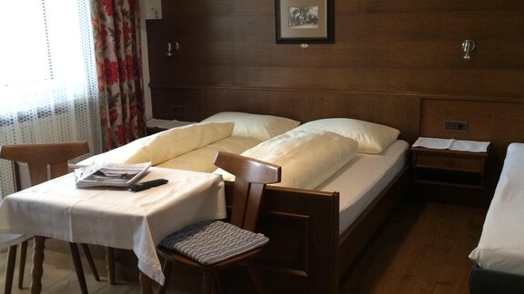 A simple room with two beds and a table. The walls are made of wood and there is a window with curtains. - Gästehaus Martin - Sölden