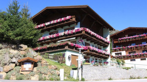A traditional alpine house with colorful flowers on the balconies. Surrounded by green trees and a clear blue sky. - Gästehaus Veit Fiegl - Sölden