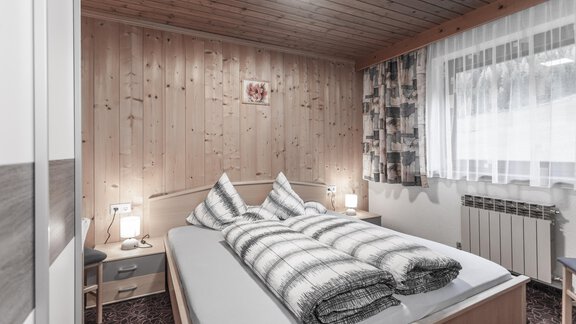 A cozy bedroom with wooden paneling and large windows. The bed is decorated with striped blankets and there is soft light from bedside lamps. - Gästeheim Schmiedhof - Sölden