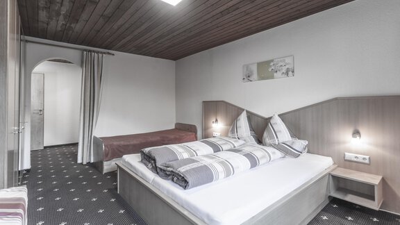 A modern bedroom with a double bed and a single bed. The walls are bright, and a cozy atmosphere is created by the wooden ceiling and soft lighting. - Gästeheim Schmiedhof - Sölden