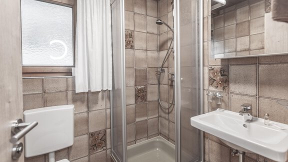 A modern bathroom with a shower, a sink, and a toilet. The walls are designed with tiles in warm tones. - Gästeheim Schmiedhof - Sölden