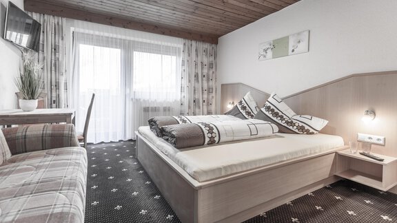 A bright bedroom with a large bed and a cozy seating area.  
Windows with curtains provide daylight and a pleasant atmosphere. - Gästeheim Schmiedhof - Sölden