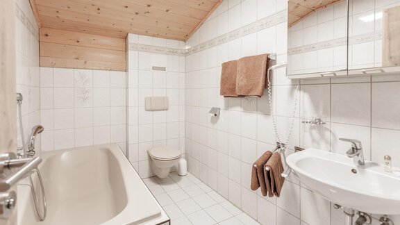 A bathroom with a bathtub, sink, and toilet. The walls are tiled and the ceiling has wood paneling. - Gästeheim Schmiedhof - Sölden