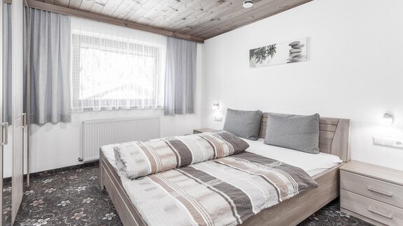 A modern bedroom with a double bed and light wood decor. The room is friendly and tidy, with large windows and soft curtains. - Gästeheim Schmiedhof - Sölden
