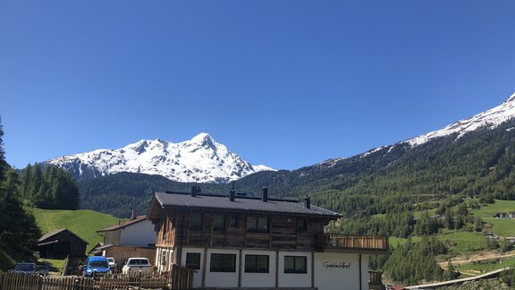 A picturesque mountain landscape with snow-capped peaks and green meadows. In the foreground stands a cozy house with balconies. - Granbichlhof - Sölden