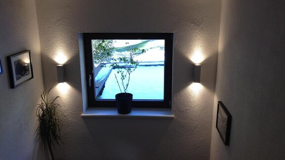 A bright stairway with wall lamps and a window. Outside, there is a view of a green area and a tree. - Haus Almrausch - Sölden