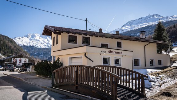 A cozy house with the inscription "Altmrausch" is located on a road in the mountains. Snow-covered mountains and a blue sky are visible in the background. - Haus Almrausch - Sölden