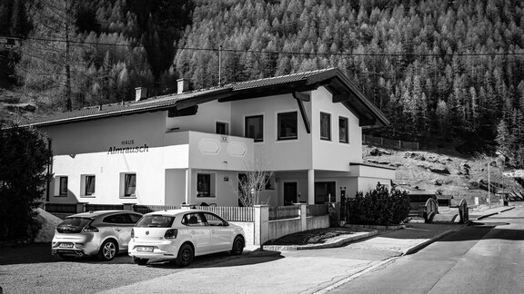 A modern building with a clear, white exterior stands in a mountainous landscape. Several cars are parked in front of the house. - Haus Almrausch - Sölden
