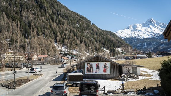 A picturesque landscape with snow-covered mountains in the background. Cars are parked along the road, and a building with advertising posters can be seen. - Haus Almrausch - Sölden