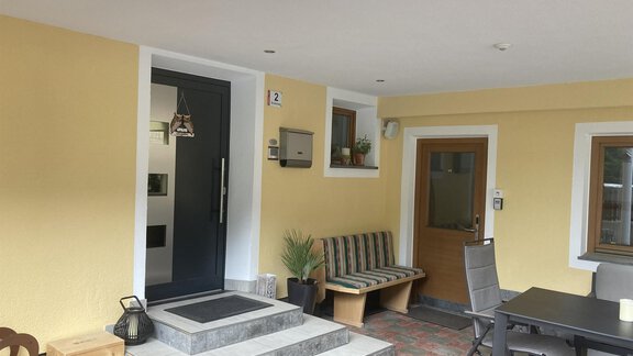 An inviting entrance area with a modern black door and a covered sitting area. The walls are painted in a friendly yellow, and there are decorative plants. - Haus Almrausch - Sölden