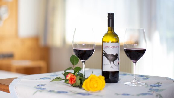 A bottle of red wine is on a table with two wine glasses. Next to it lies a rose and a yellow flower on a doily. - Haus Alpenstern - Sölden