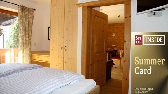 A cozy bedroom with wooden paneling and a large window. In the background, a living room is visible, and there is a display for the Ötztal Summer Card. - HAUS ERIKA apartments - Sölden