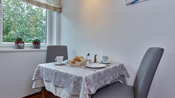A small table with a white tablecloth, set with plates, cups, and a breakfast. In the background, plants and a window with a view of greenery can be seen. - Haus Fontana - Sölden