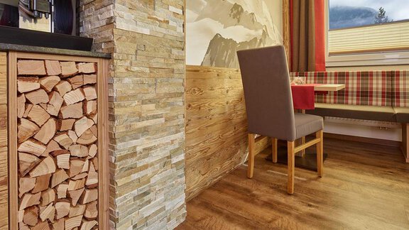 A cozy room with wooden paneling and a stone wall. In the background, there is a table with benches and a window with a view of the mountains. - Haus Fridolin - Sölden