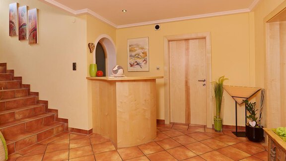 A bright hallway with a staircase and a reception desk. The walls are painted in warm colors, and there are plants in the room. - Haus Fridolin - Sölden