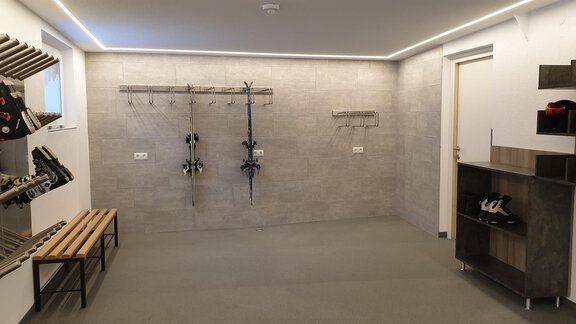 A modern room with gray walls and a light floor. Ski equipment holders are hanging on the wall, and there is a bench as well as a shelf. - Haus Fridolin - Sölden