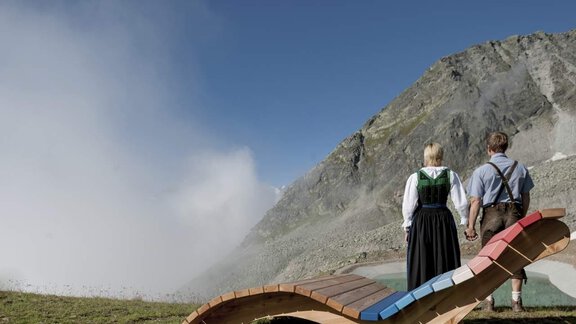 A couple in traditional clothing stands on a wooden deck and looks at an impressive mountain landscape. In the background, fog and high mountains are visible. - Haus Montjola - Sölden