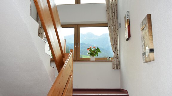 A stairwell with bright walls and a wooden railing. Through the window, you can see mountains and a flower plant is on the windowsill. - Haus Romana - Sölden