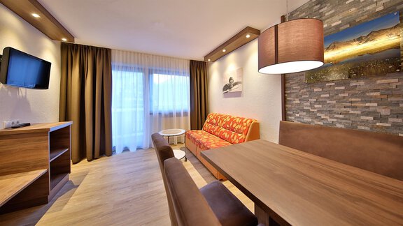 A stylish living room with an orange sofa, a wooden table, and modern furniture. Large windows let in plenty of light and create an inviting atmosphere. - Haus Söldenblick - Sölden
