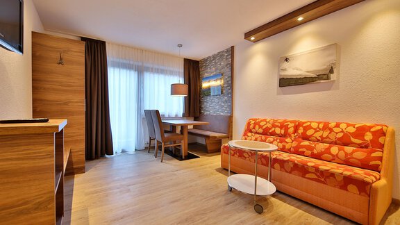 A modern living room with an orange couch and a small table. Large windows let in plenty of light and provide a view outside. - Haus Söldenblick - Sölden