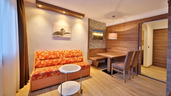 A cozy living room with an orange couch and a small table. In the background, there is a dining table with chairs and soft light. - Haus Söldenblick - Sölden