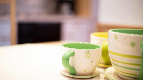 A white table with colorful cups in shades of green and yellow. In the background, a modern kitchen is visible. - Haus Sonnwend - Sölden