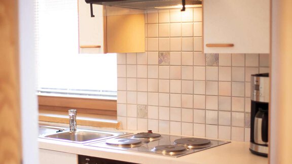 A modern kitchen with a stove, sink, and coffee machine. The walls are tiled with light-colored tiles and there is plenty of natural light. - Haus Sonnwend - Sölden