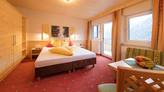 A comfortable hotel room with a large bed that is colorfully decorated. Next to the bed is a small table with fresh fruits and a window with a view outside. - Haus Stefanie - Sölden