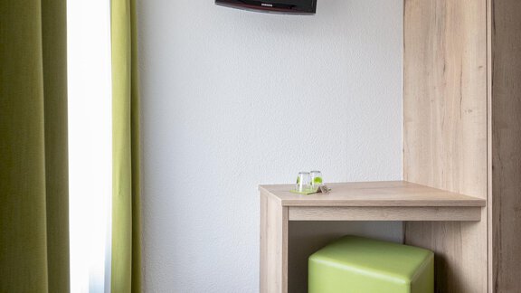 An interior with a desk and a green stool. A television is hanging on the wall, and the windows are decorated with green curtains. - Haus Ulrich - Sölden