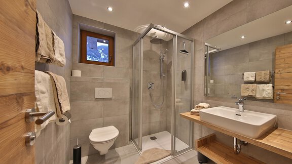 A modern bathroom with a glass shower and a large mirror. The walls are in neutral gray tones, and there are wooden accents. - Haus Söldenblick - Sölden