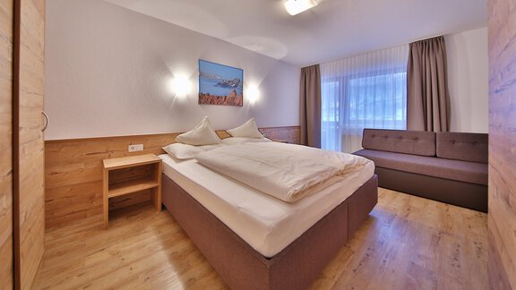 A modern bedroom with a large bed and a cozy sofa corner. The large windows let in plenty of light, and the wooden details create a warm atmosphere. - Haus Söldenblick - Sölden