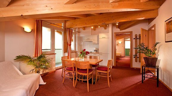 A bright apartment with wooden ceilings and a red carpet. A dining table with chairs and an open kitchen are clearly visible. - Hotel Garni Ehrenreich - Sölden