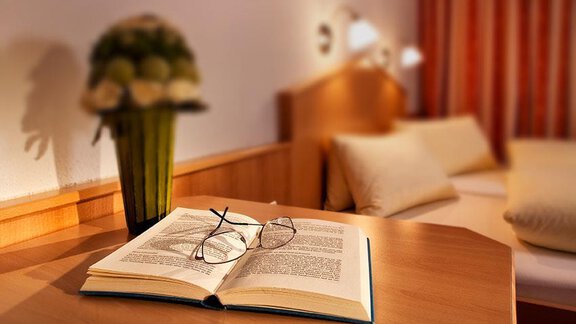 An open book lies on a table, next to it are glasses and a flower vase. In the background, a sleeping area and soft light can be seen. - Hotel Garni Ehrenreich - Sölden