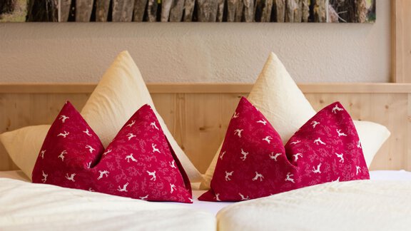 Two decorative pillows in the shape of mountains are lying on a bed. The pillows are red with a pattern and are placed on a light bed cover. - Hotelappart Peter - Sölden | © Falkner Georg