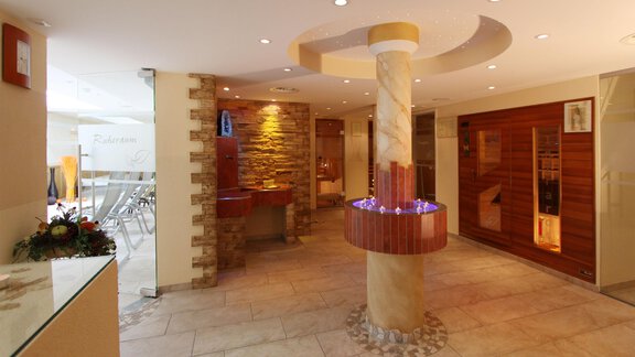 An elegant wellness area with a stylish reception desk and a decorative column fountain. In the background, saunas and cozy seating options can be seen. - Hotelappart Peter - Sölden | © Falkner Georg