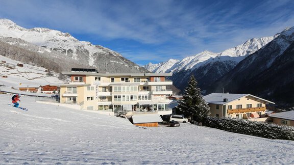 A snow-covered landscape with a modern building and the surrounding mountains. Skiers are using the slopes nearby. - Hotelappart Peter - Sölden